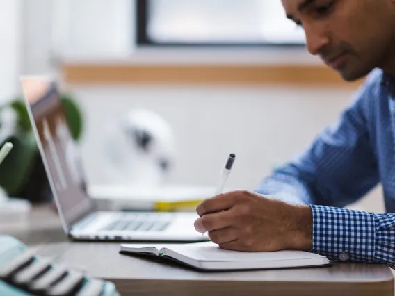 Man writing in notebook next to open laptop